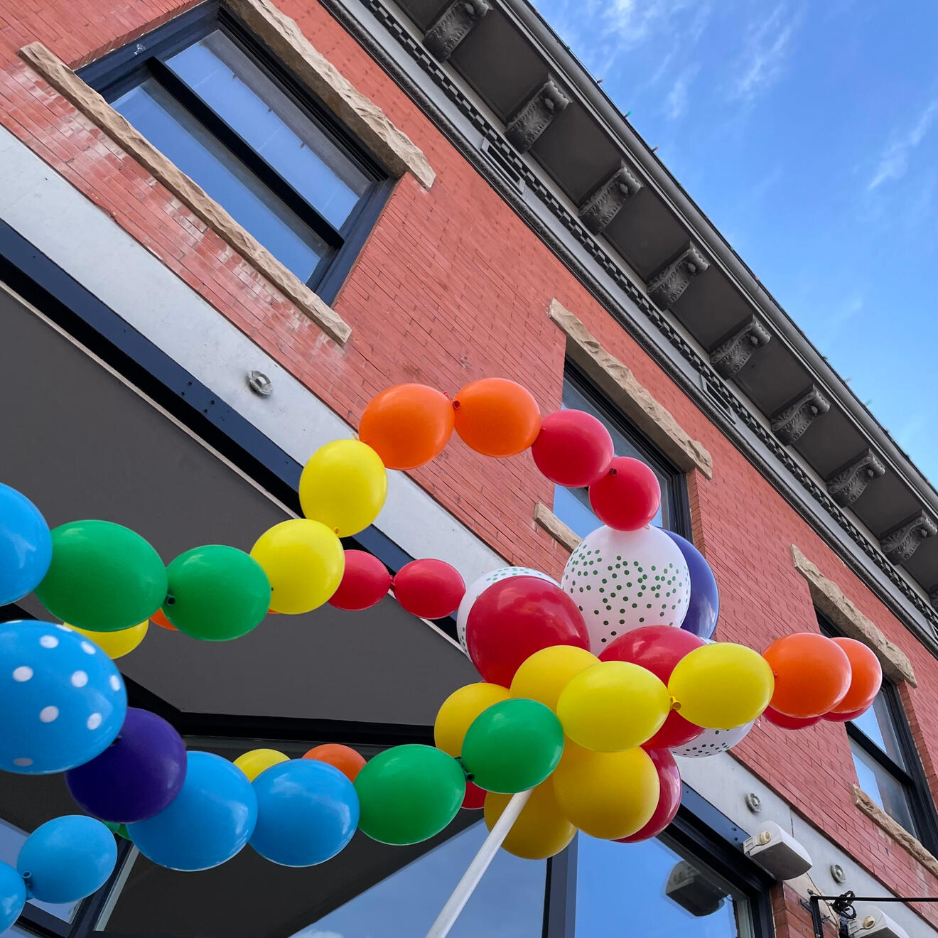 Low-angle photograph of a bundle of balloons in all colors of the rainbow. The camera looks up at the balloons, which float in the air in front of the red brick facade of a building in Fort Collins' Old Town Square.
